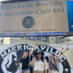 Three women smiling in front of a sign that reads "Marrickville Bowling and Recreation Club".